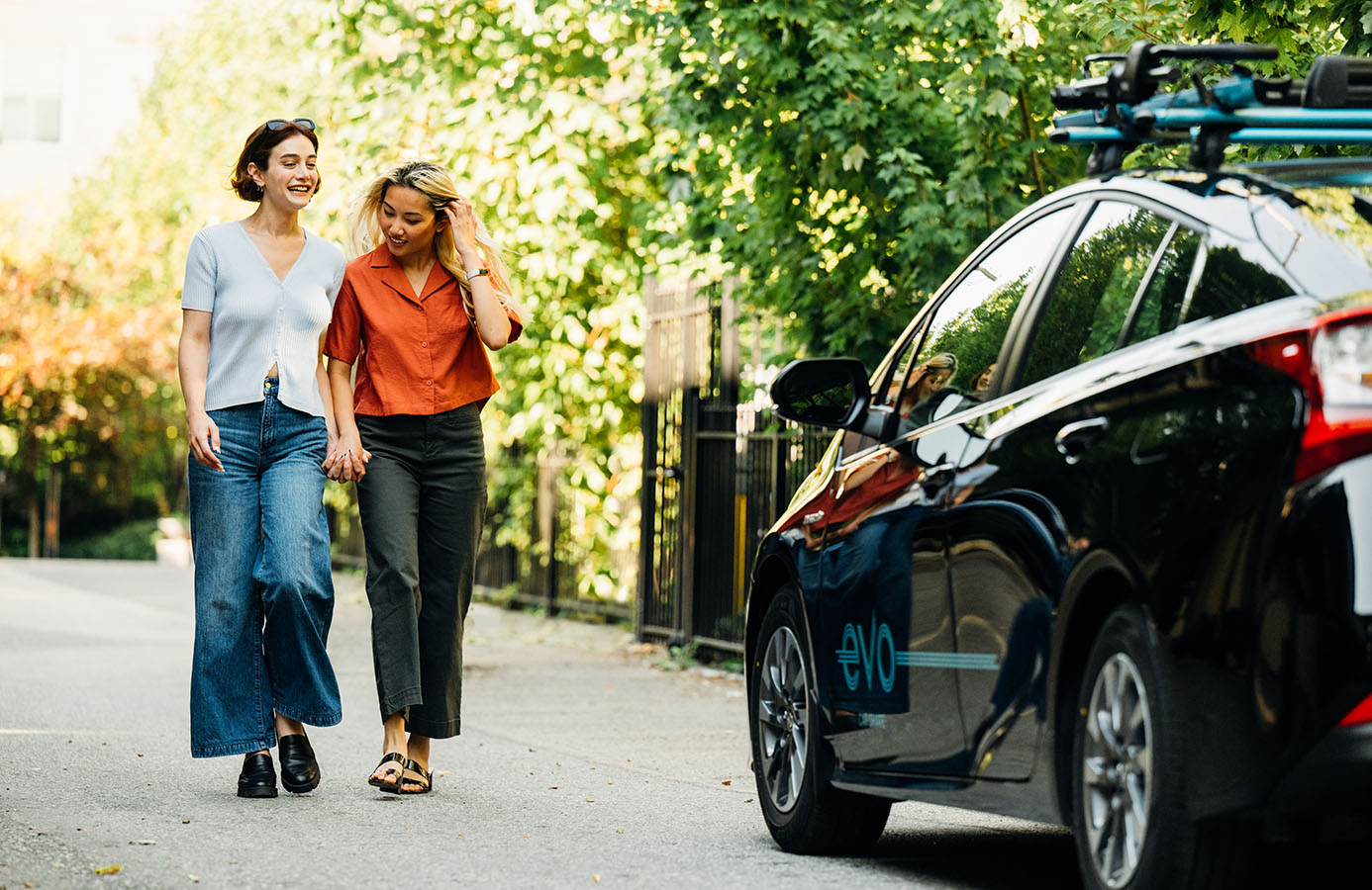 Two women holding hands walking alongside a parked Evo